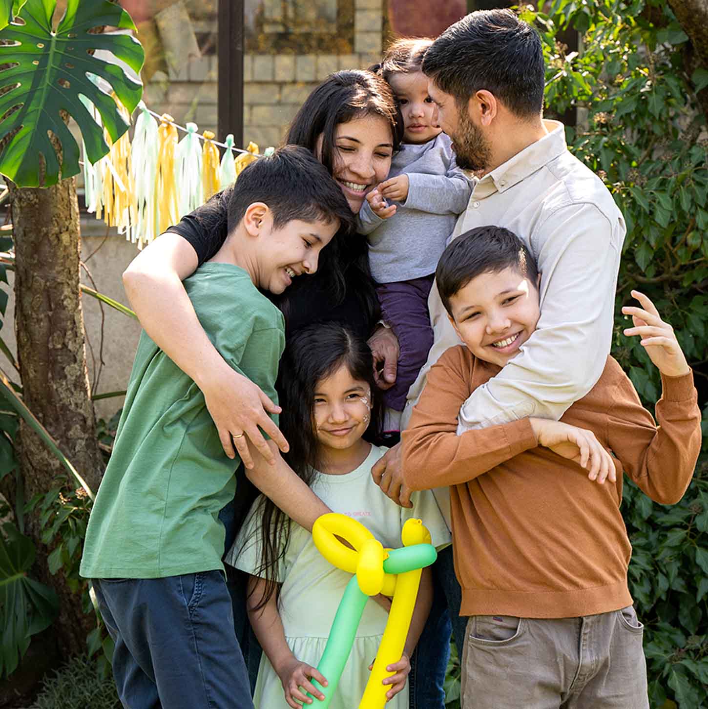 Familia mexicana de seis integrantes, tres hombres y tres mujeres, los padres tienen alrededor de 38 años y los niños entre 6 a 13 años, visten de manera casual y están abrazados muy felices en el patio de su hogar.